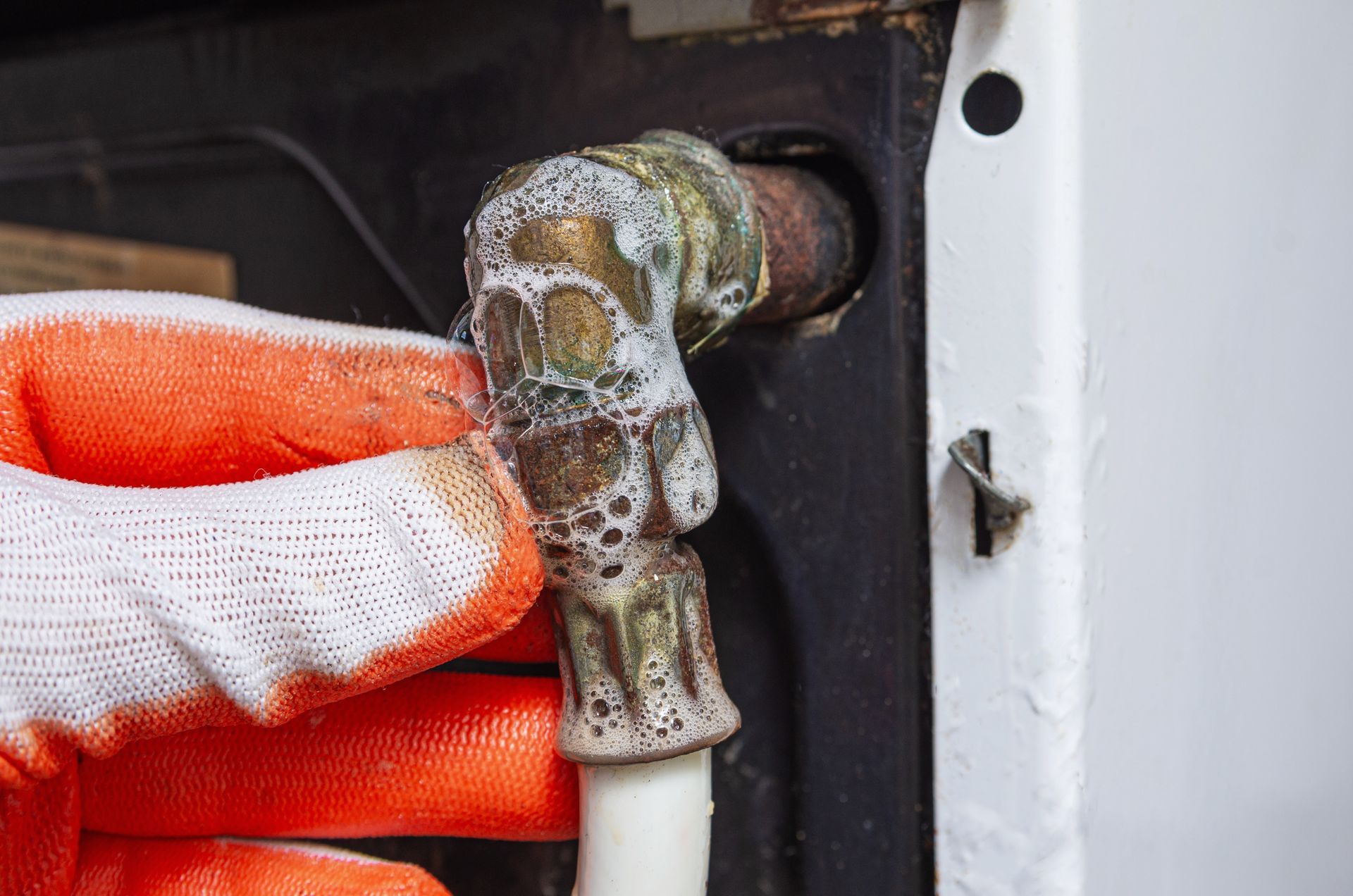A gloved hand holds a corroded, calcified brass pipe fitting connected to a white water hose on an appliance.