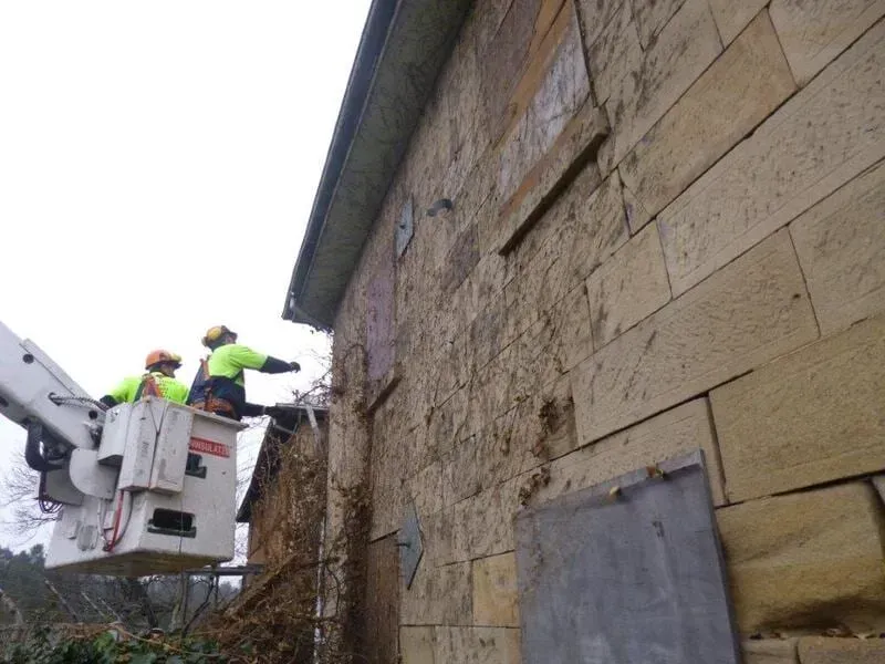Two workers in a lift bucket inspect a stone building. They wear hard hats and high-vis vests. Overcast sky.