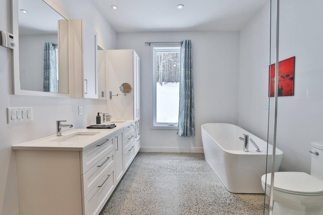 Salle de bains blanche avec baignoire îlot, grande vasque et fenêtre avec rideau bleu.