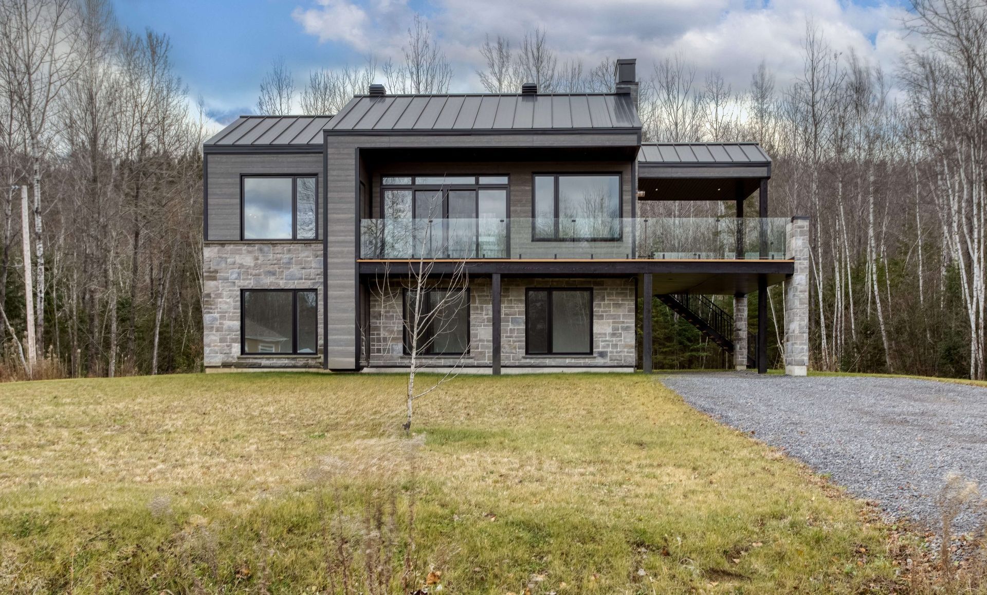 Maison moderne à deux étages avec toit en métal gris, façade en pierre et balcon donnant sur une allée de gravier et un jardin verdoyant.