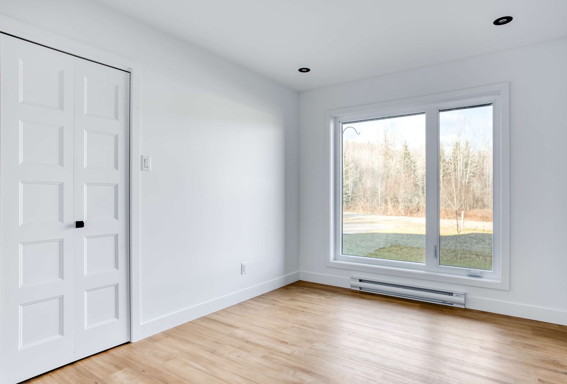 Pièce blanche vide avec porte fermée, fenêtre donnant sur l'extérieur et parquet.