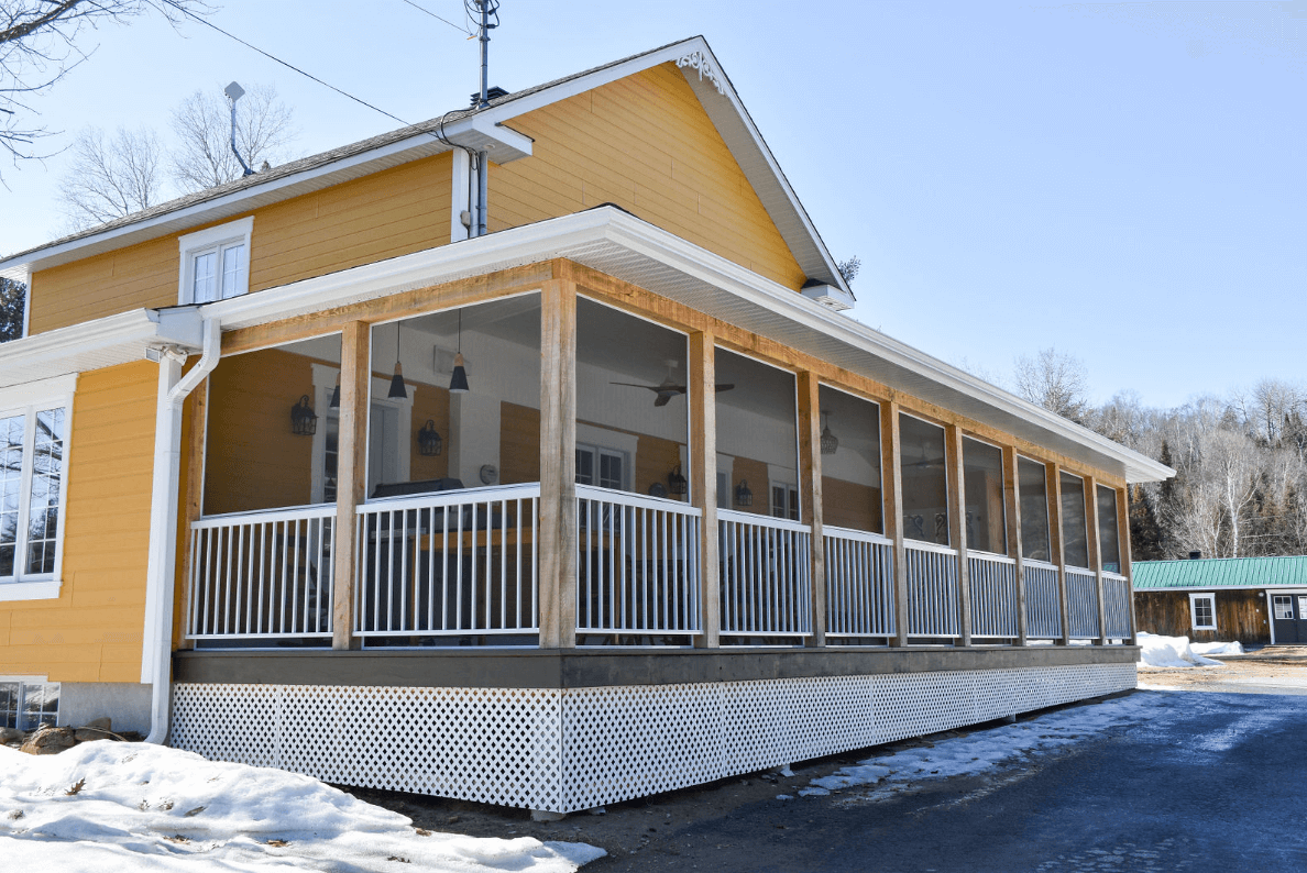 Maison jaune avec une véranda grillagée sur une terrasse surélevée ; neige au sol.
