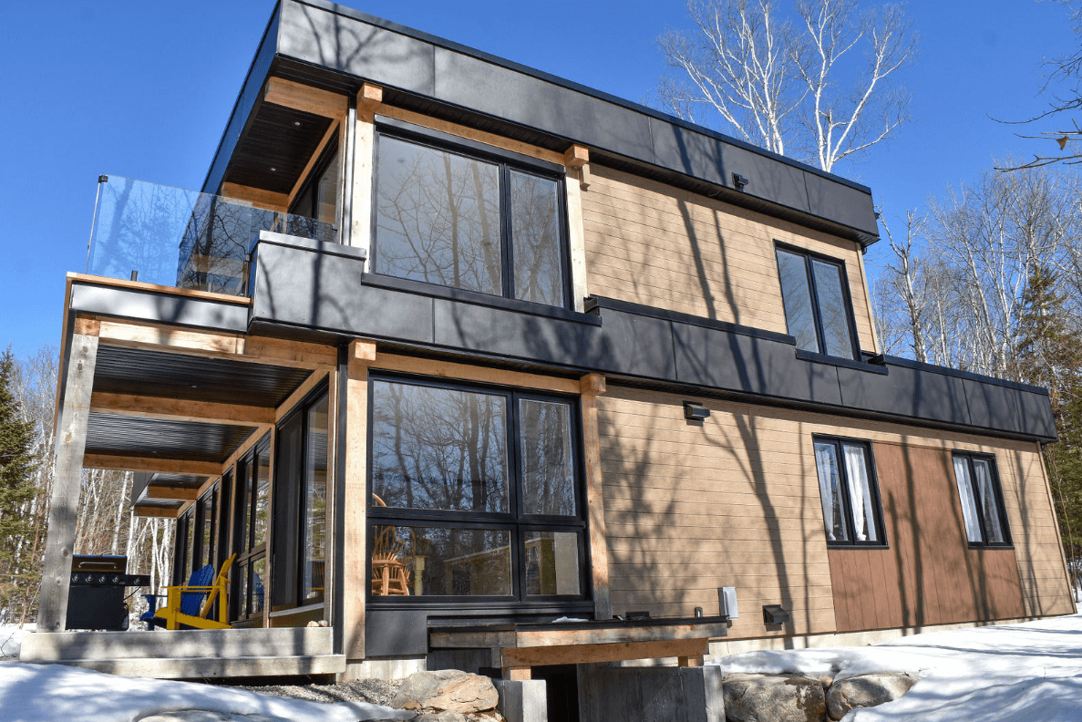 Maison moderne à deux étages avec de grandes fenêtres, des éléments en bois et un balcon, située dans une zone boisée et enneigée.