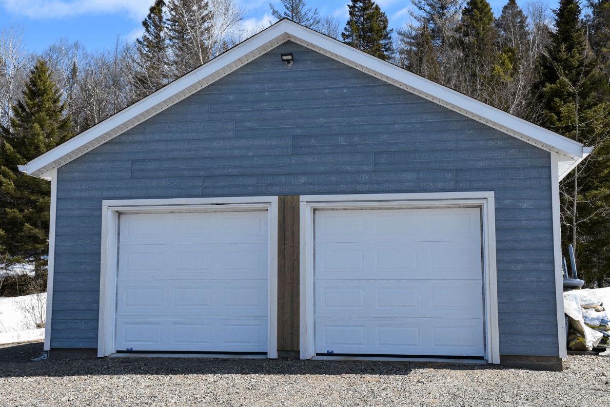 Garage bleu pour deux voitures avec portes blanches, se détachant sur un fond d'arbres et de ciel lumineux.