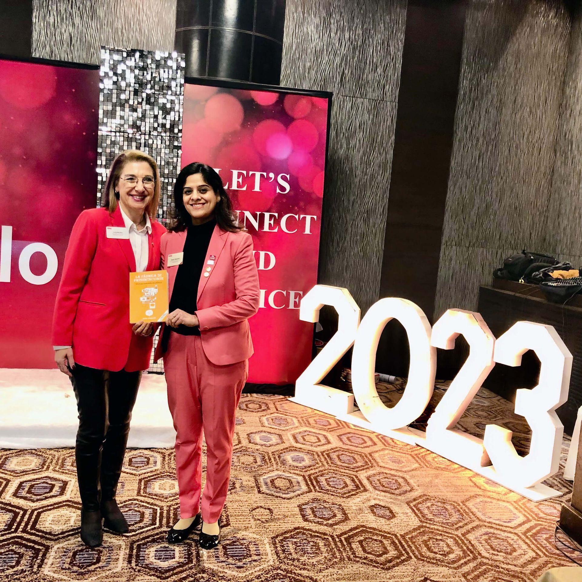 Dos mujeres posan juntas. Una de rojo y otra de rosa, sosteniendo un libro frente a un cartel de 2023 en un evento.