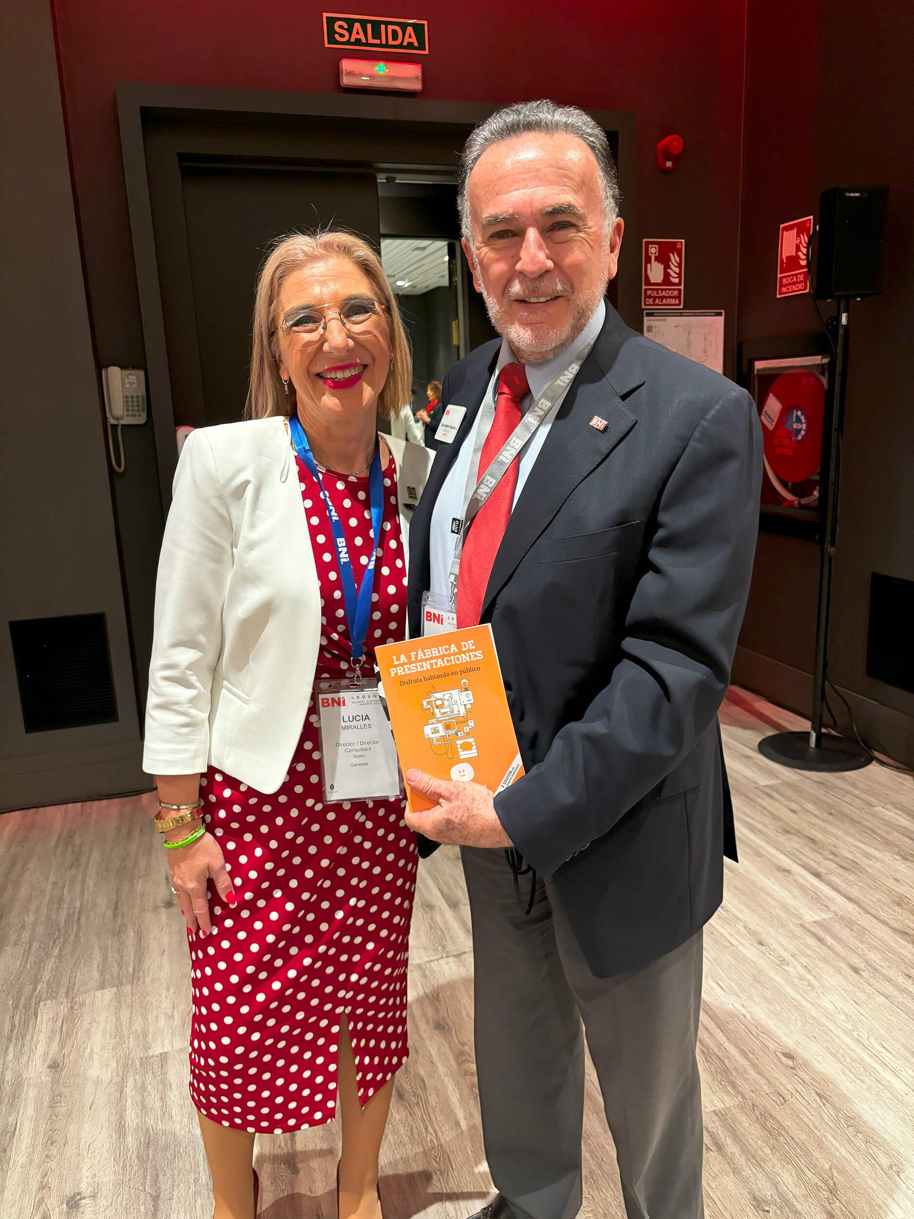 Dos personas sonriendo, posando juntas en un interior. Una mujer con un vestido rojo de lunares y un hombre con traje sosteniendo un libro.
