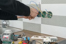 Hand wiring electrical boxes in a wall, with tools on a counter below.