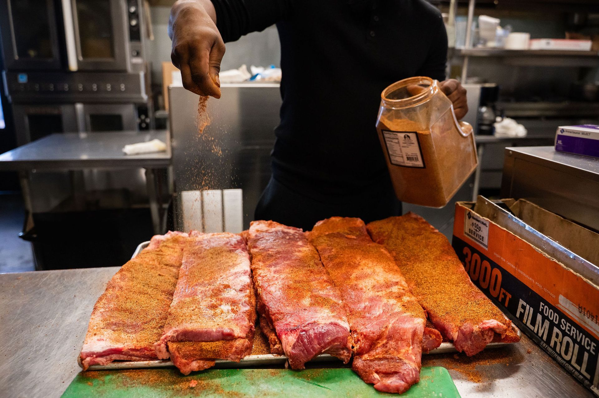 Ribs being seasoned on a tray in a kitchen, with a hand holding a spice jar nearby