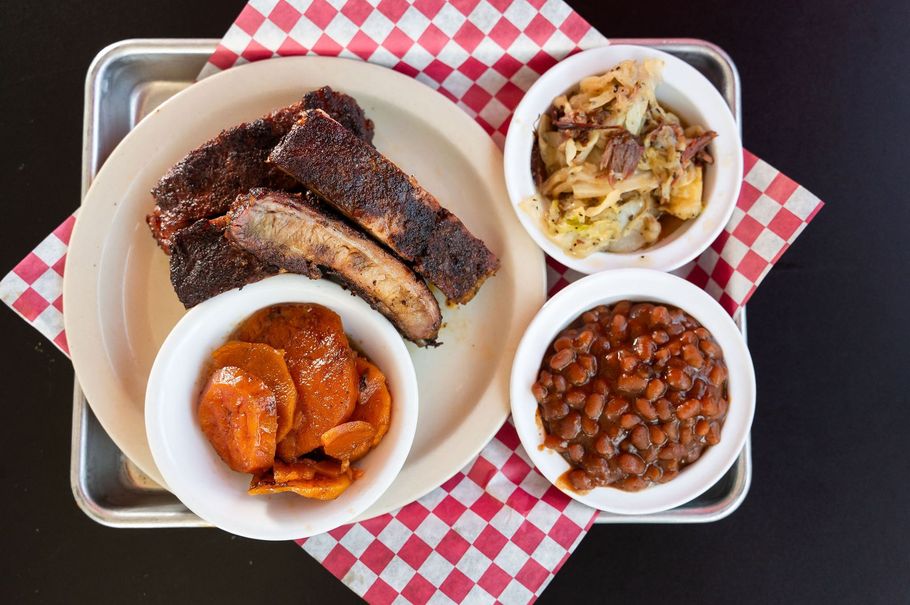 Tray of barbecue ribs, baked beans, potato salad, and sweet potatoes on a red checkered paper liner.