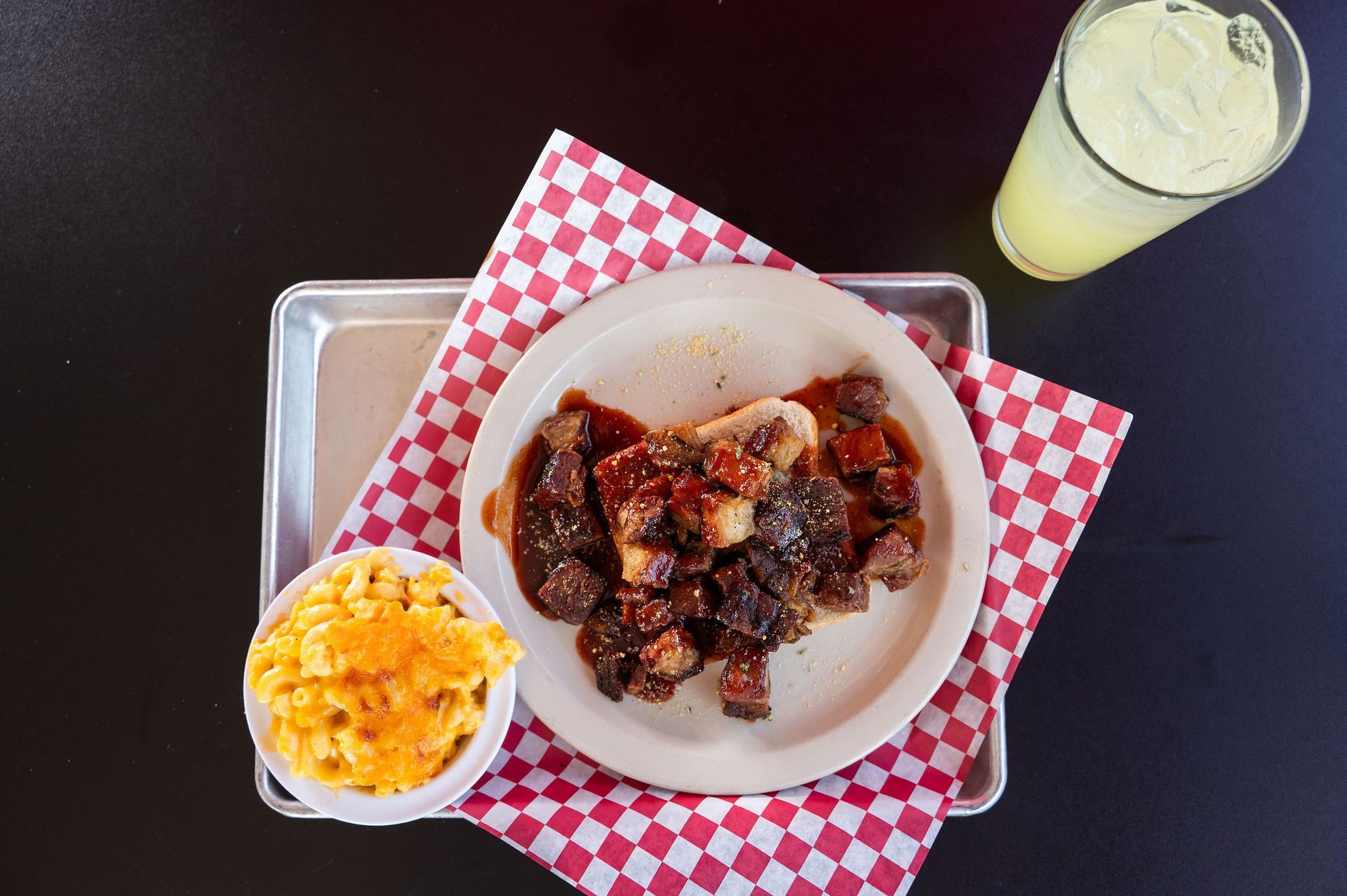 Plate of saucy ribs with macaroni and cheese, plus a lemonade on a tray with a red checkered napkin.
