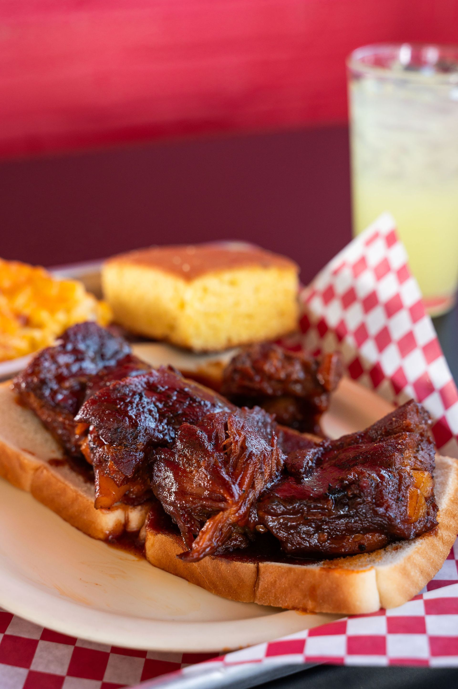 Barbecue ribs on a tray with cornbread, coleslaw, and a lemonade drink on a red-checkered tablecloth