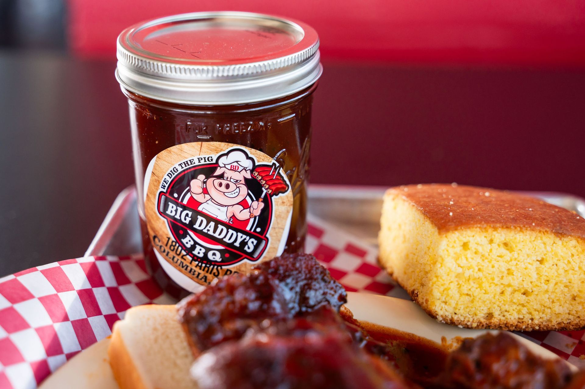 Jar of BBQ sauce on a red-checkered tray beside cornbread and ribs.