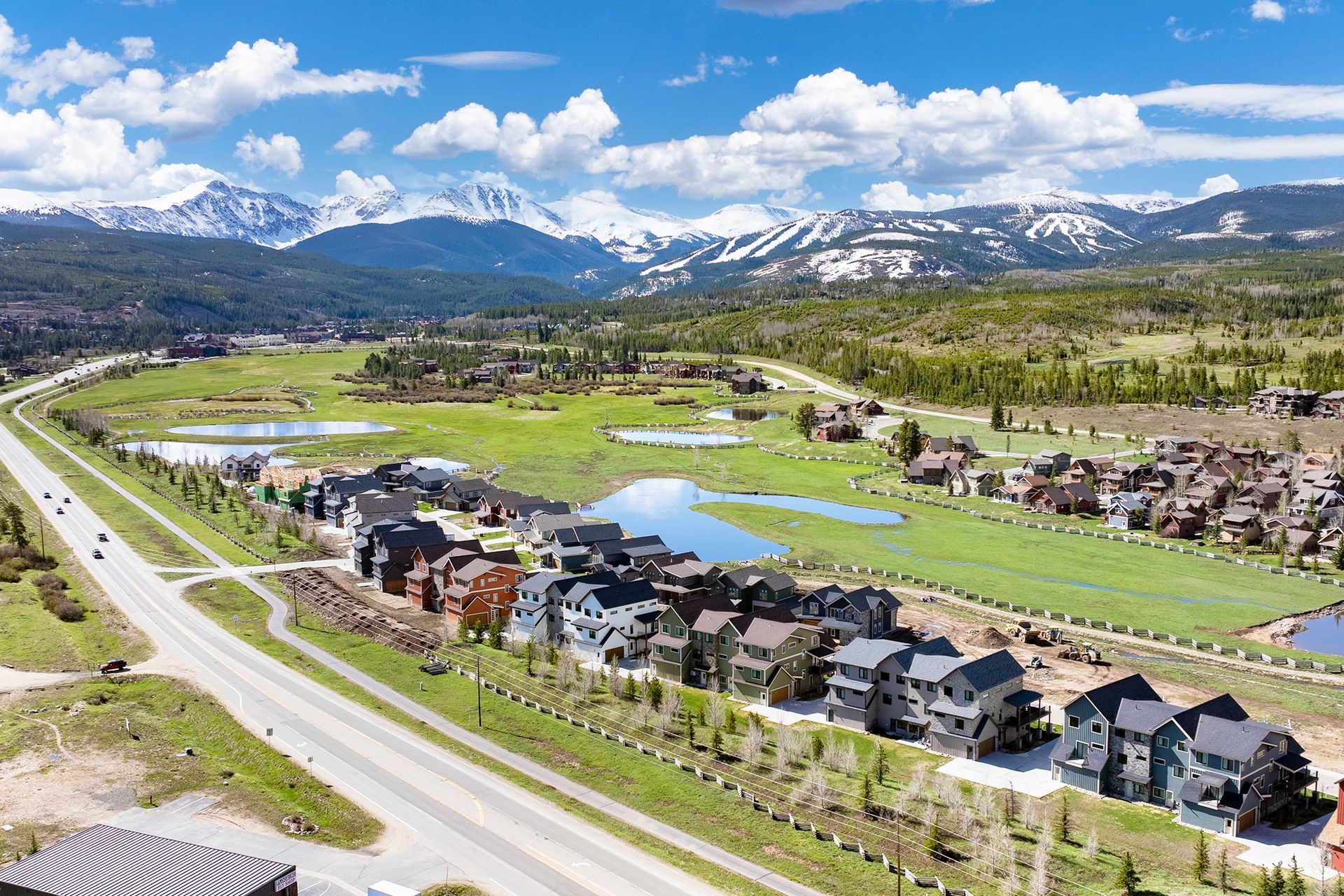 Houses next to a road with a green field, ponds, and mountains in the background under a blue sky.