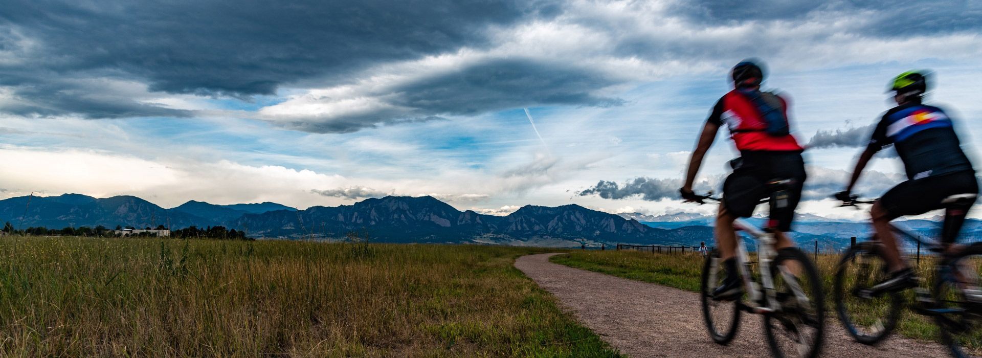 Two cyclists ride mountain bikes down a dirt path with mountains in the background. Cloudy sky.