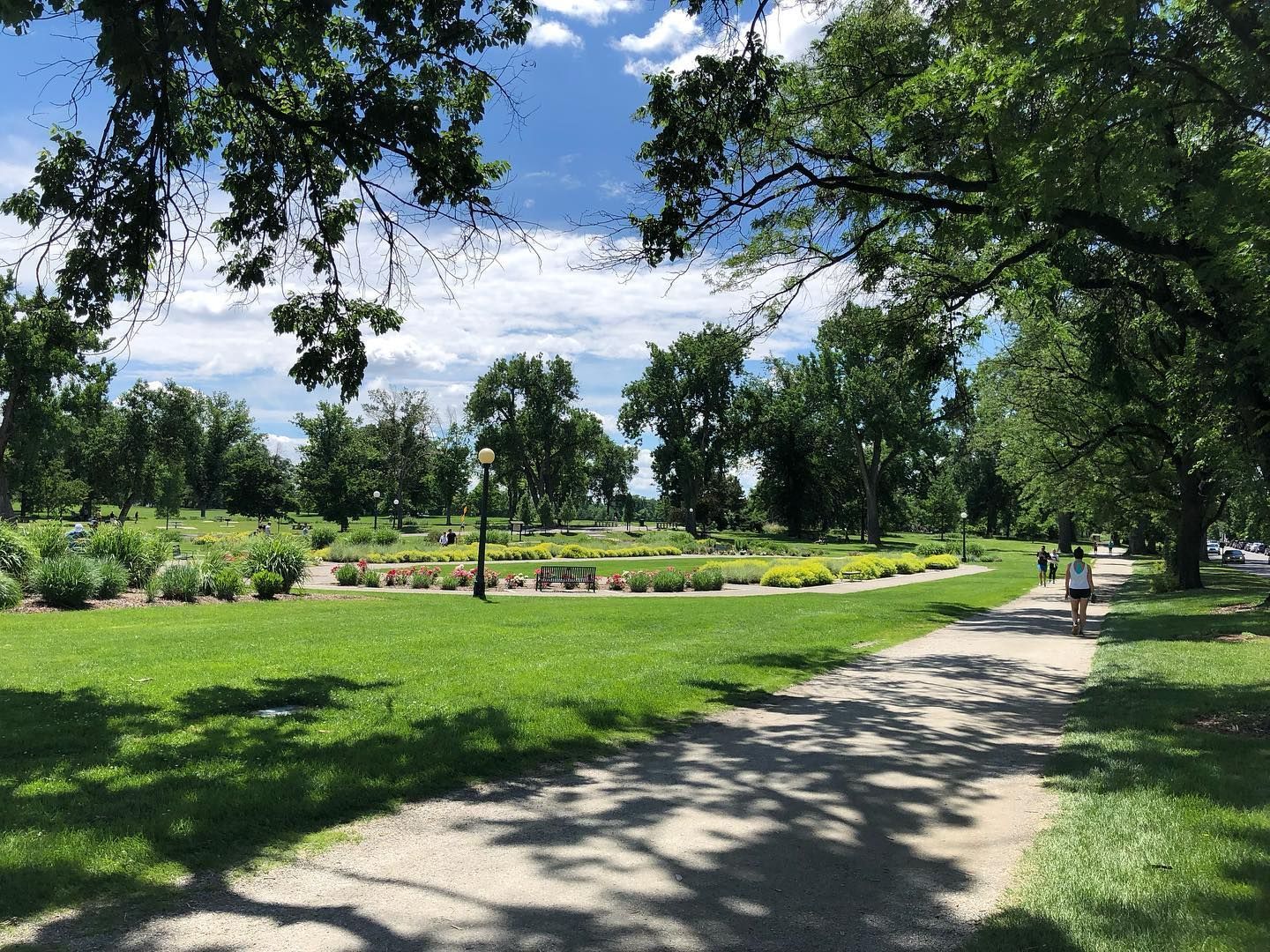 Park scene with grassy areas, walking path, trees, flowers, and a person walking.