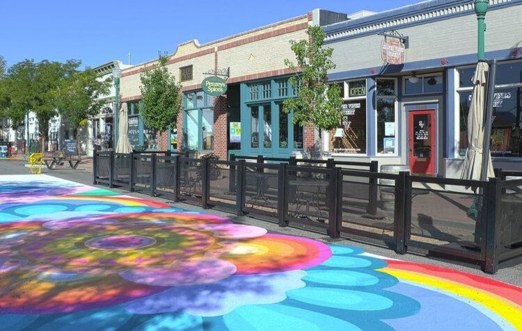 Colorful street mural in front of shops with outdoor seating. Sunny day with blue sky.