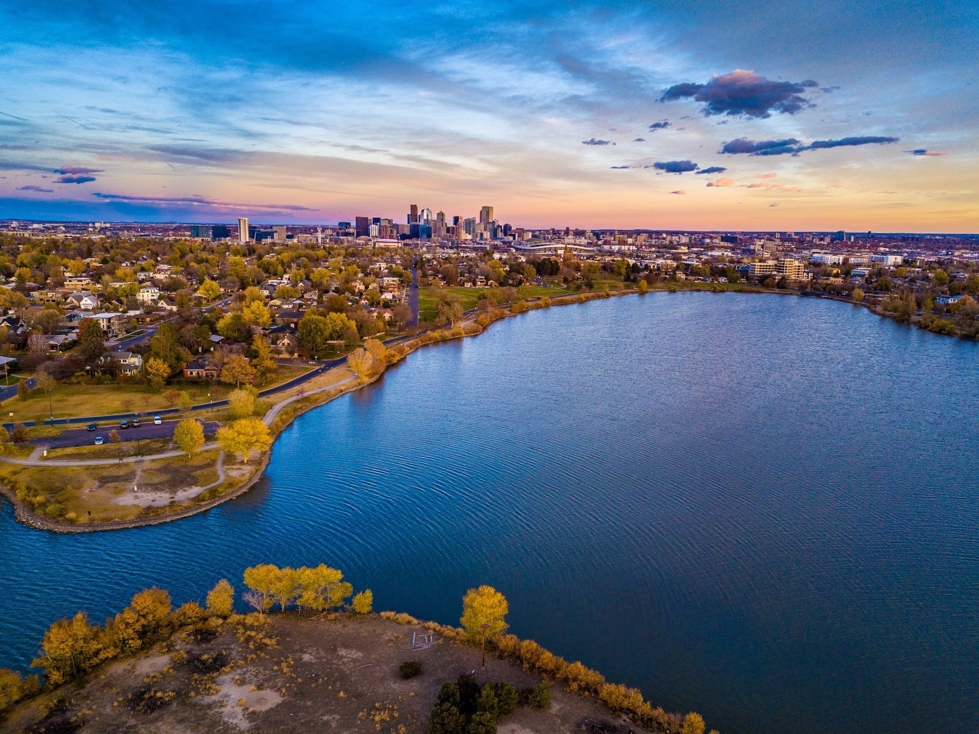 Aerial view of a lake with a city skyline in the distance, under a colorful sunset.