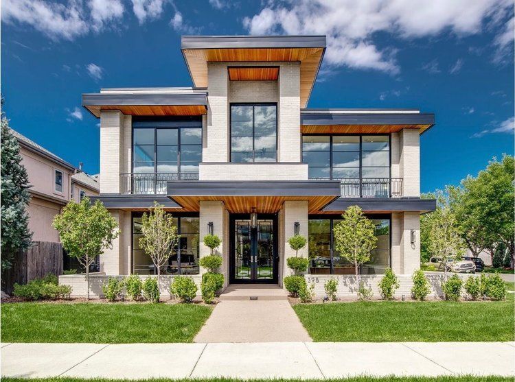 Modern two-story house with large windows, light stone facade, wooden accents, and manicured lawn under a blue sky.