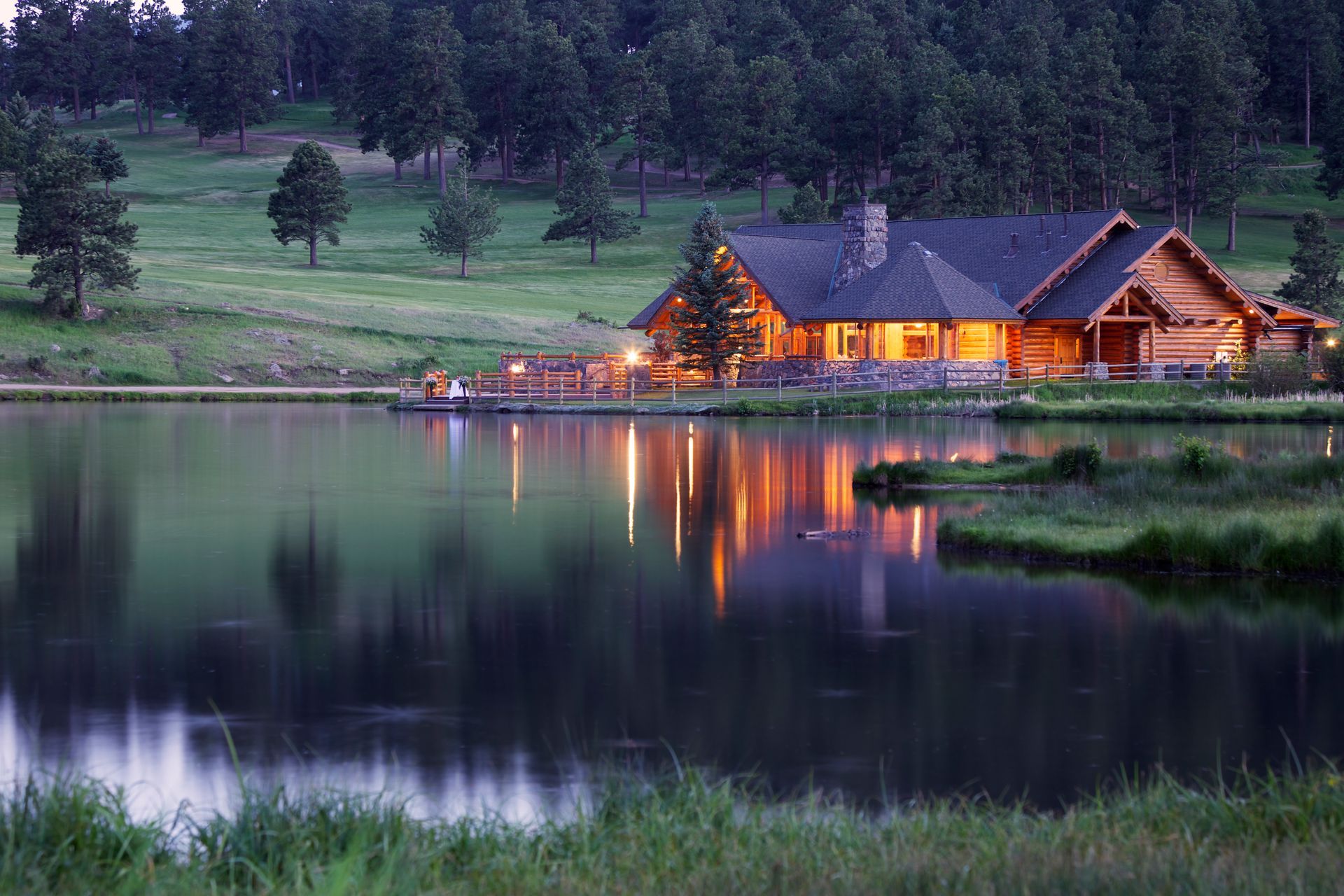Lakeside log cabin at dusk, lights reflecting on the water. Lush green grass and trees in the background.