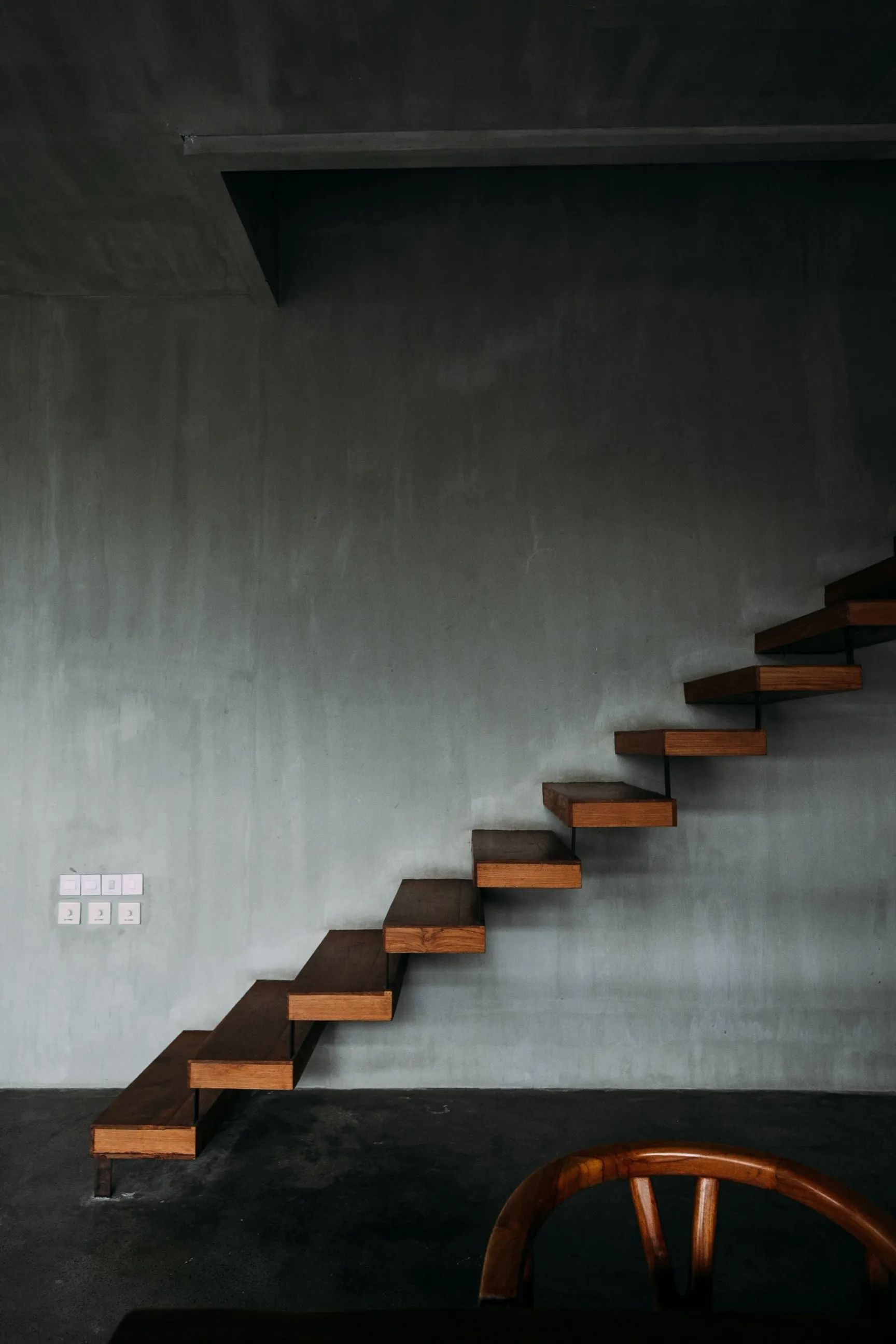 Floating wooden stairs against a gray wall, with a curved wooden chair in the foreground.