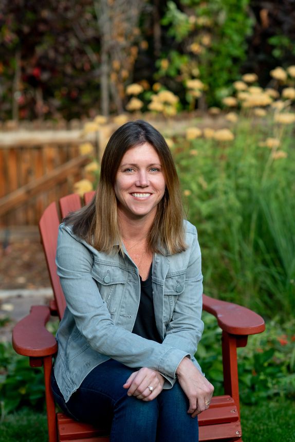 Woman in jeans and jacket sits in a red chair outdoors, smiling at camera.