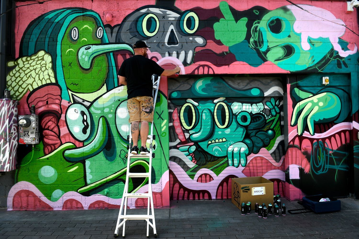 Man on ladder paints colorful mural with cartoon figures.