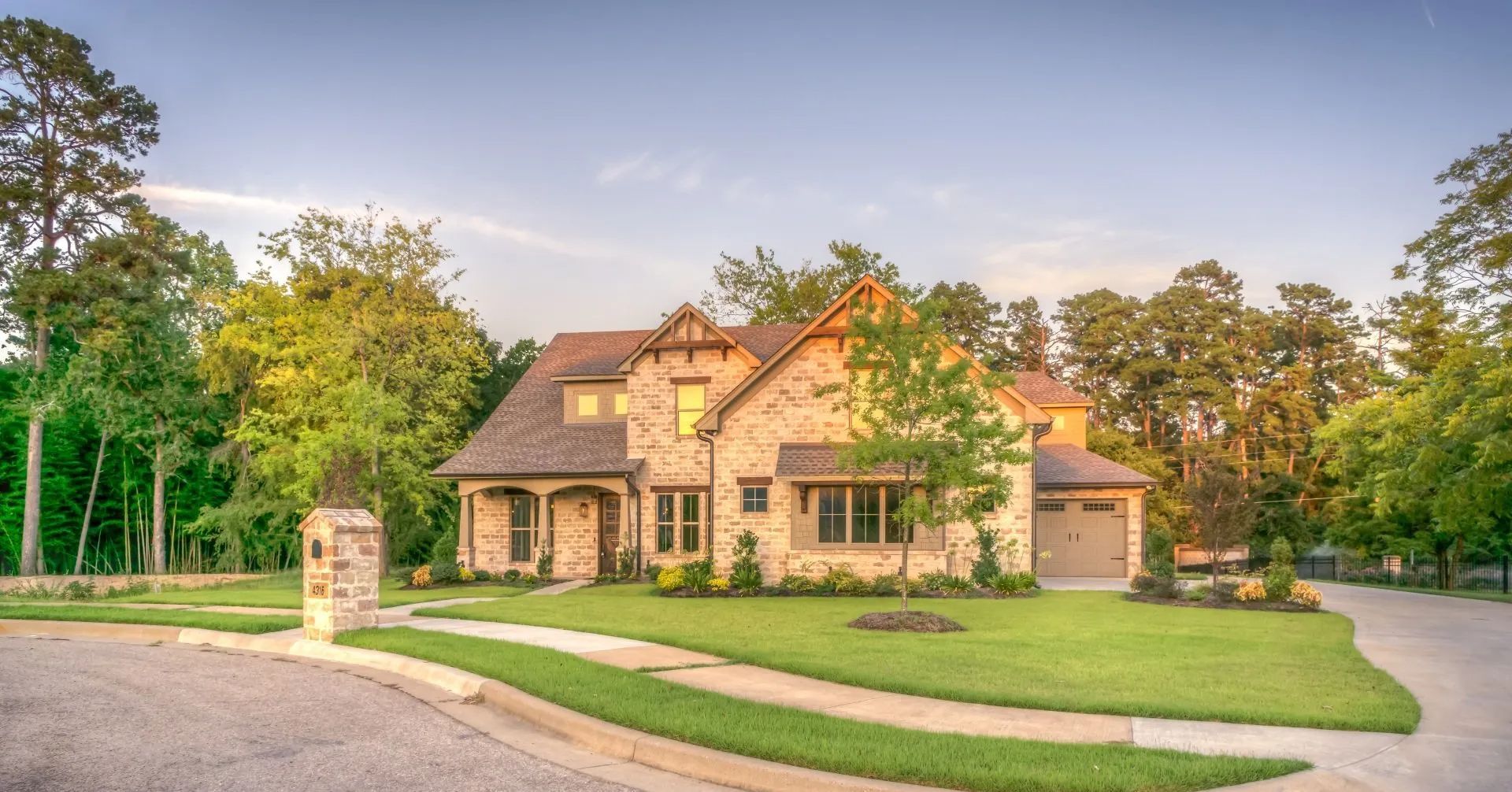 Two-story brick house with green lawn, driveway, and trees under a blue sky.