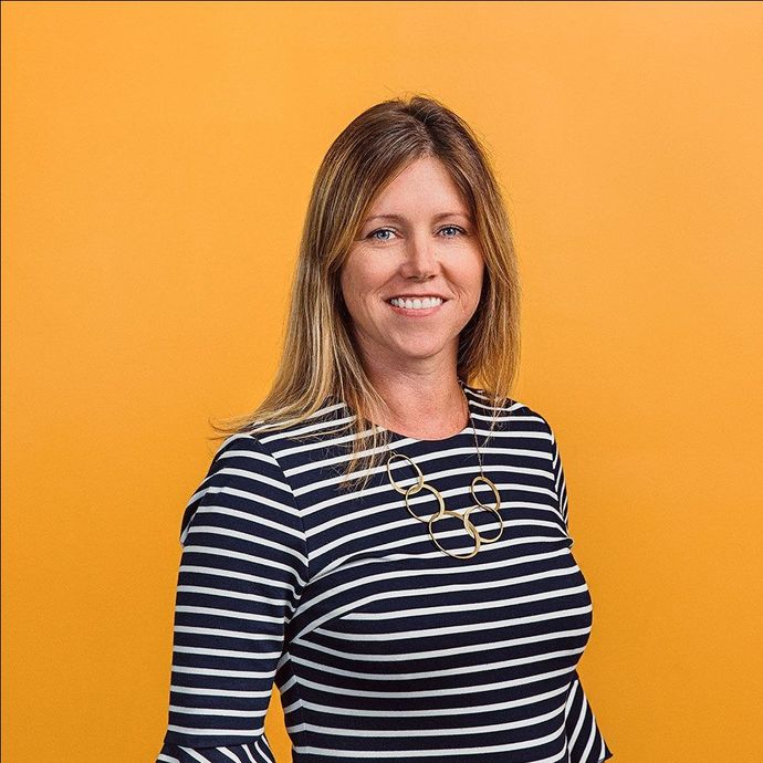 Woman with blonde hair smiles, wearing a striped shirt and gold necklace against an orange background.