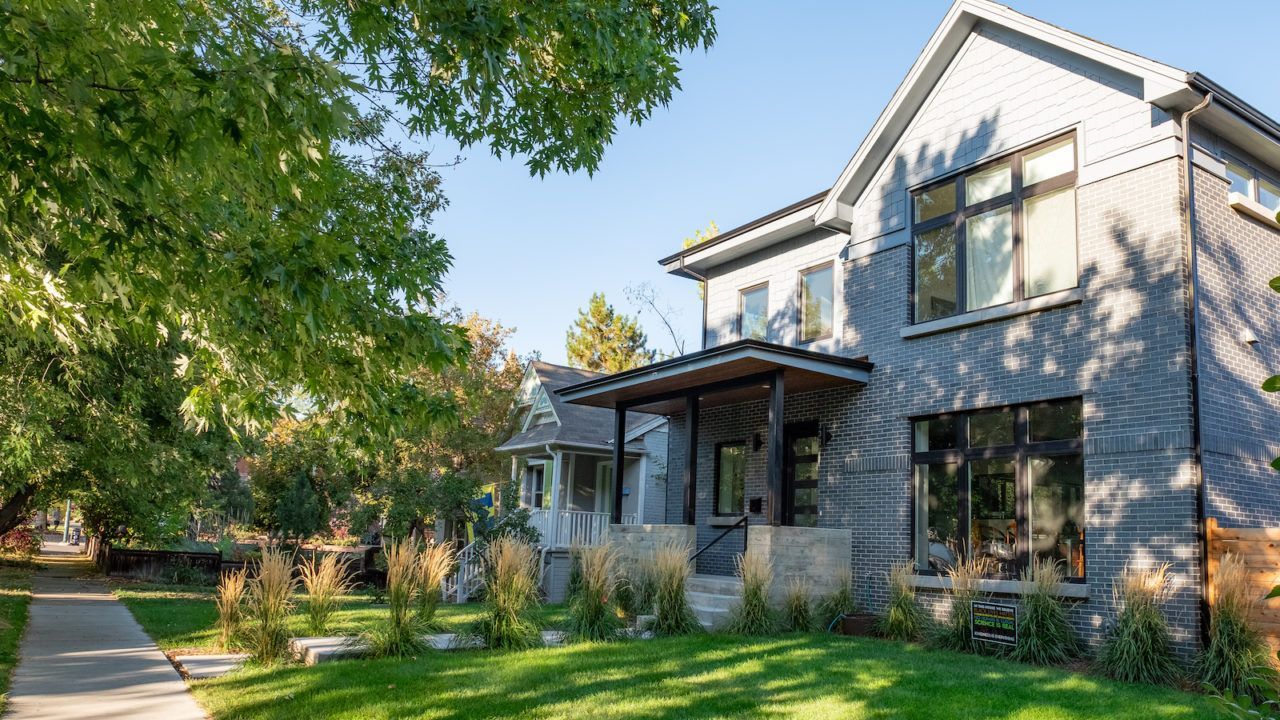 Modern two-story home with blue-grey siding and large windows. Green lawn, sidewalk, and trees in a sunny setting.