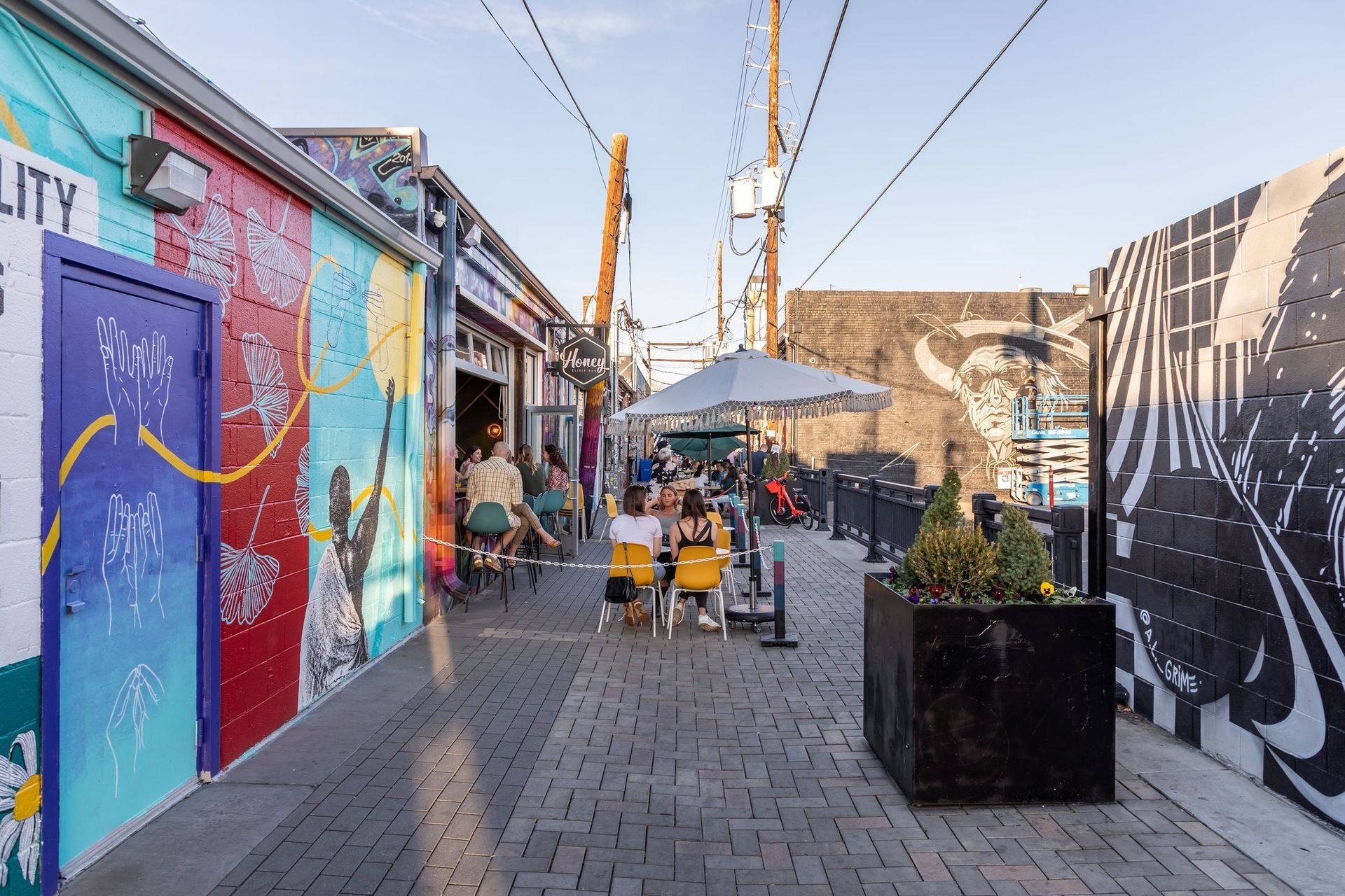 Outdoor dining alley with colorful murals, people seated at tables under an umbrella.