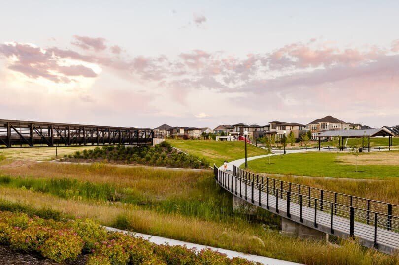 Wooden bridges and pathways over a grassy area, leading to houses under a sunset sky.