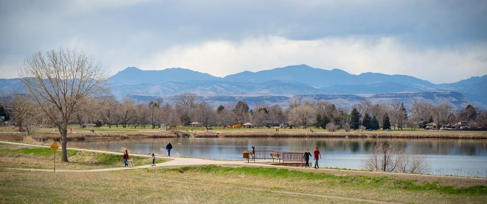 A lake surrounded by trees and mountains under a cloudy sky. People are on a path near the water.