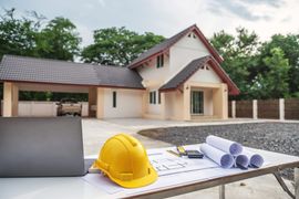 A yellow hard hat, blueprints, and a laptop sit on a table in front of a house under construction.