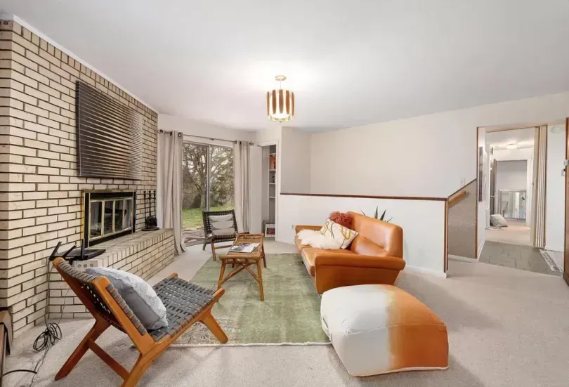 Living room with brick fireplace, orange couch, and wooden chair, looking out to a yard.