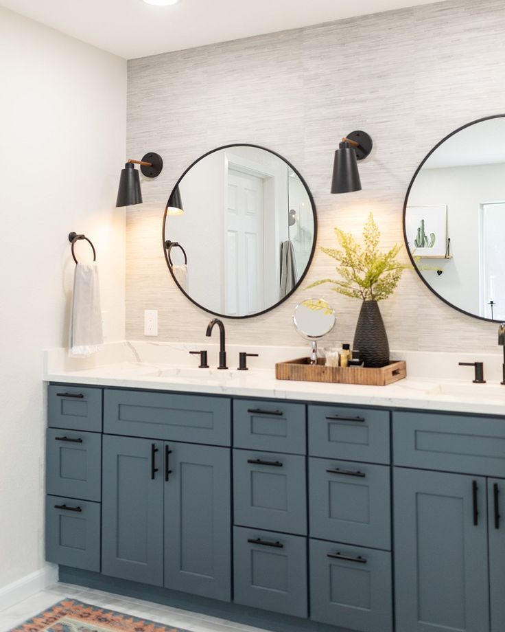 Blue bathroom vanity with two round mirrors, black fixtures, sconces, and a vase with greenery.