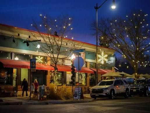 Nighttime storefront with red awnings, lit holiday decorations, pedestrians, and a parked SUV