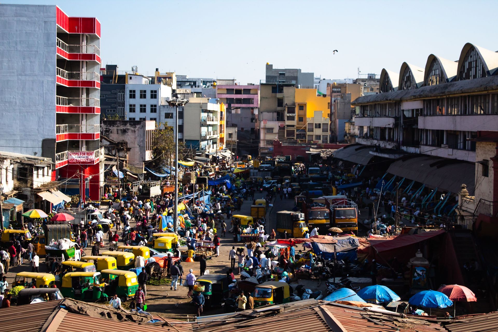 Busy urban market street with crowds, colorful taxis, and multi-story buildings under a clear sky
