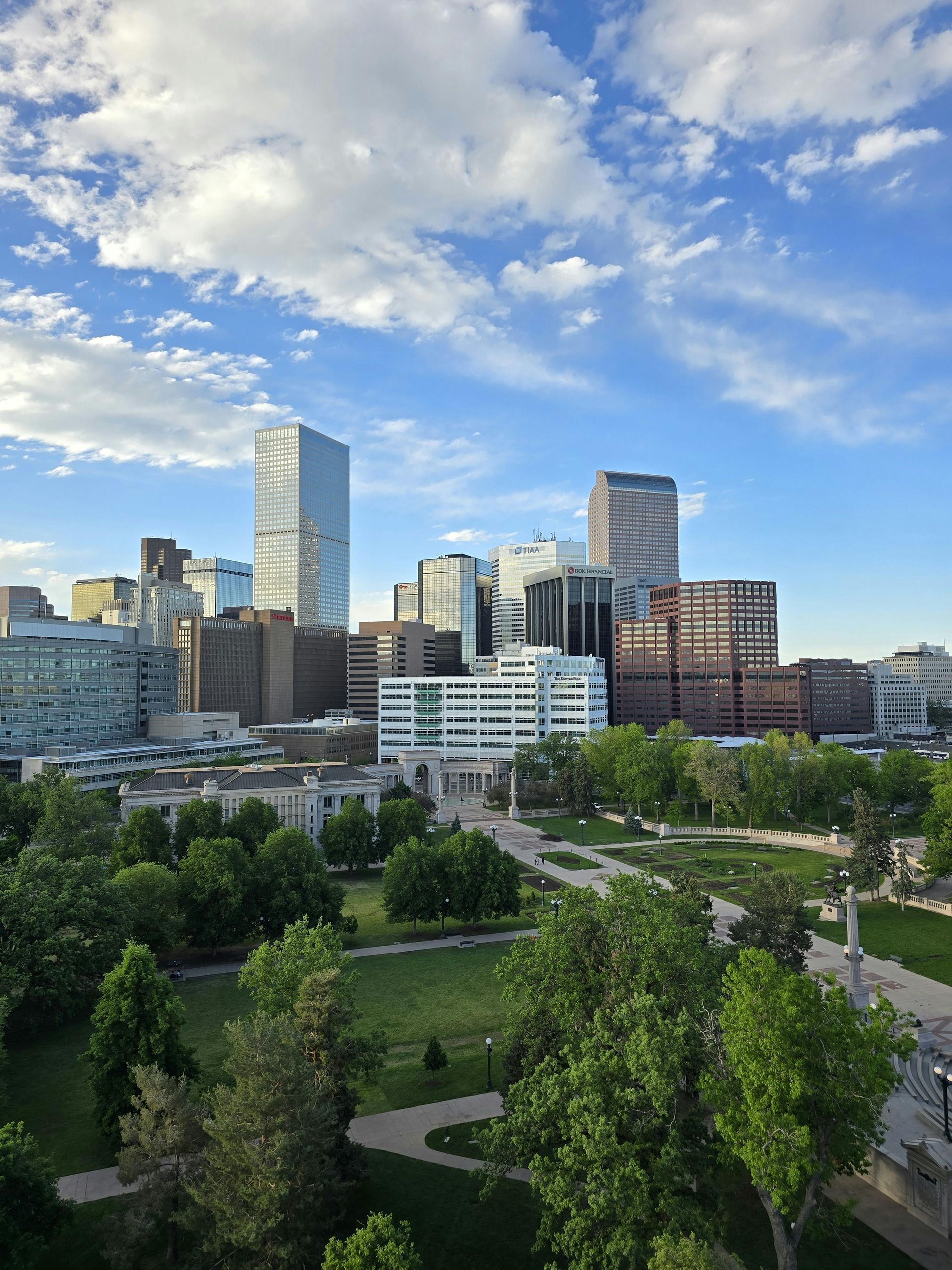 City skyline with green trees in a park below a blue sky with fluffy clouds.