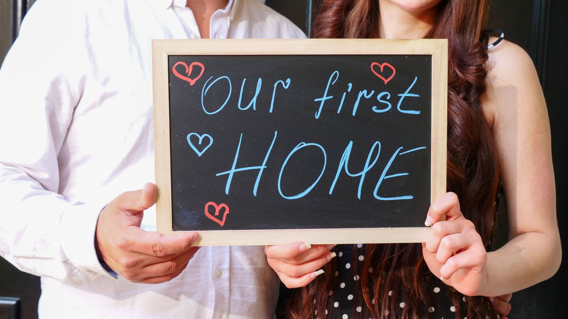 Couple holding chalkboard sign reading “Our first home” with small hearts
