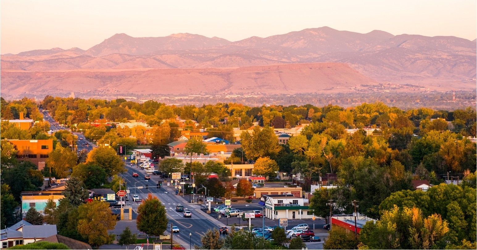 Town with buildings and trees, road with cars, mountains in the background, lit by warm sunset colors.