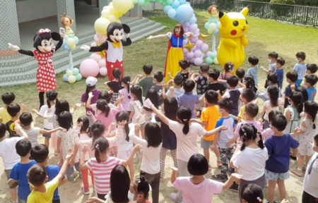 A group of children are standing in a circle with balloons and mascots.