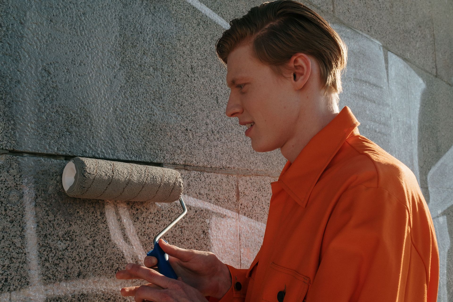 Person in orange work clothes paints a gray brick wall with a roller outdoors.