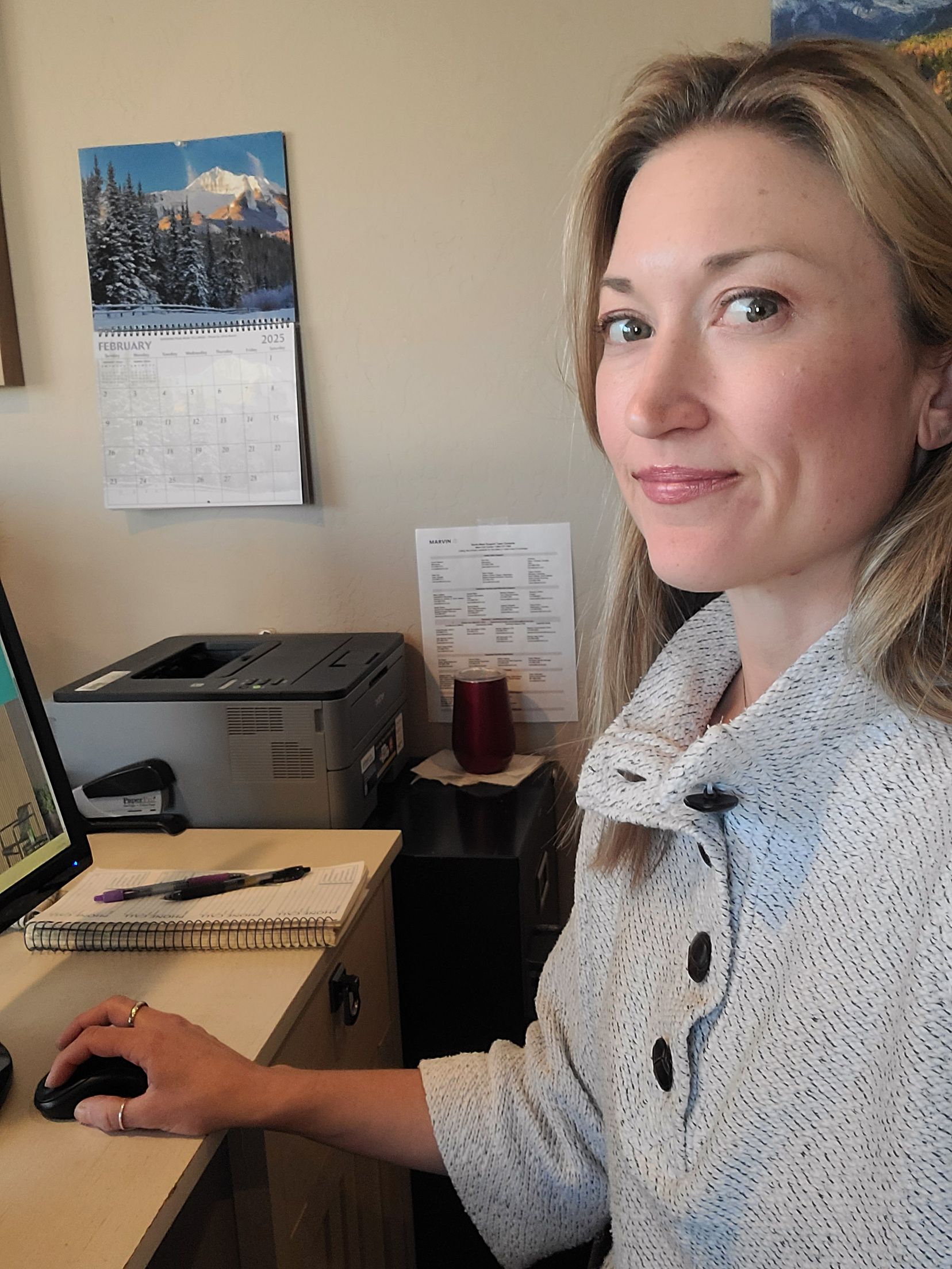 A woman is sitting at a desk with a computer and a calendar on the wall behind her