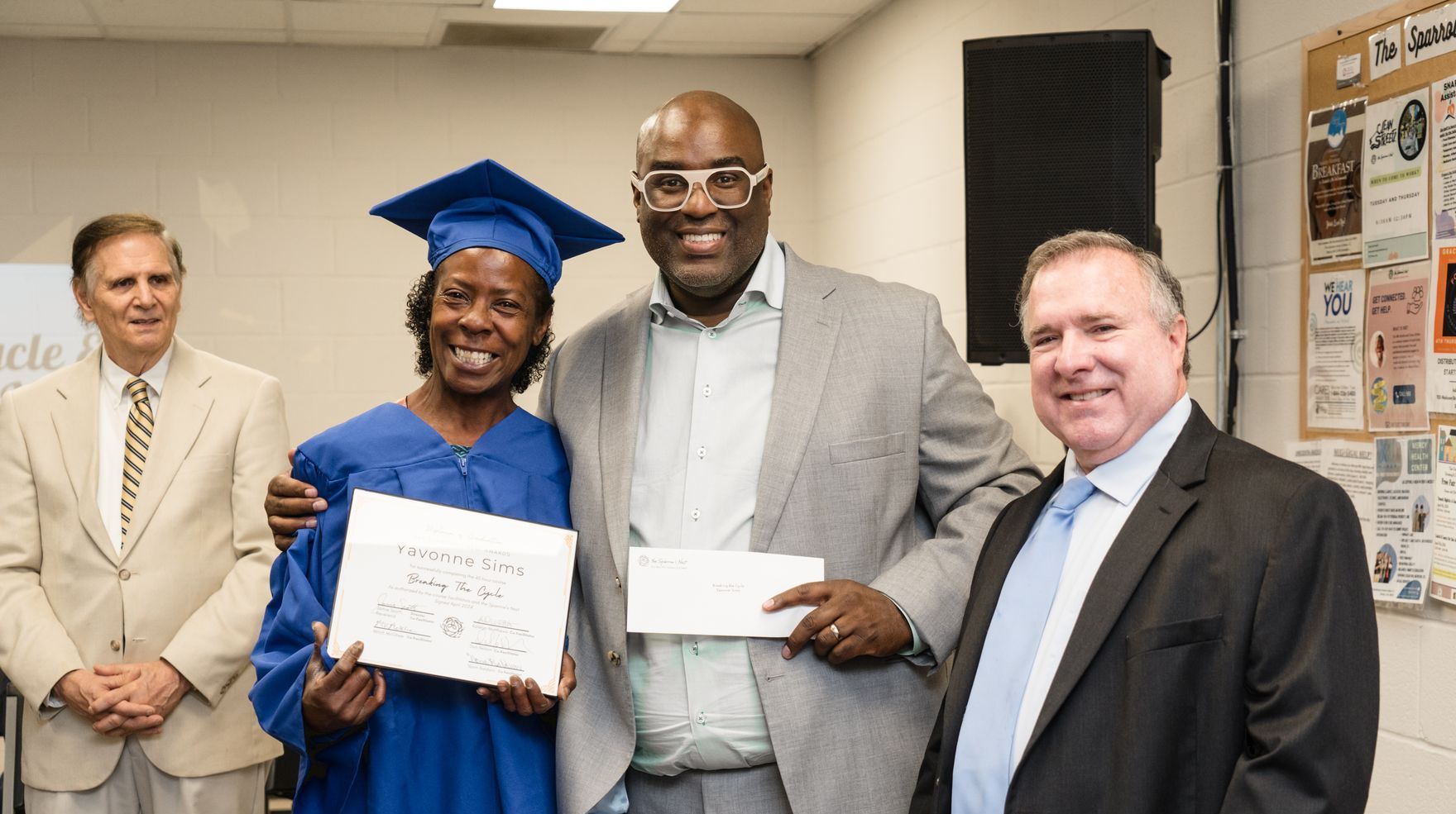 Graduate in blue cap and gown holds diploma beside two smiling men and another man in office setting