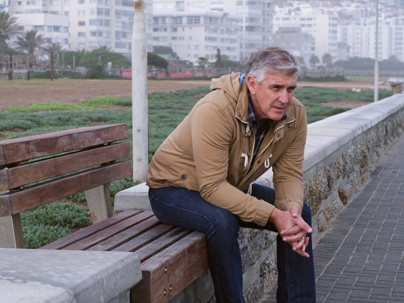 Man in tan jacket sits on a bench, looking downcast near a coastal walkway.