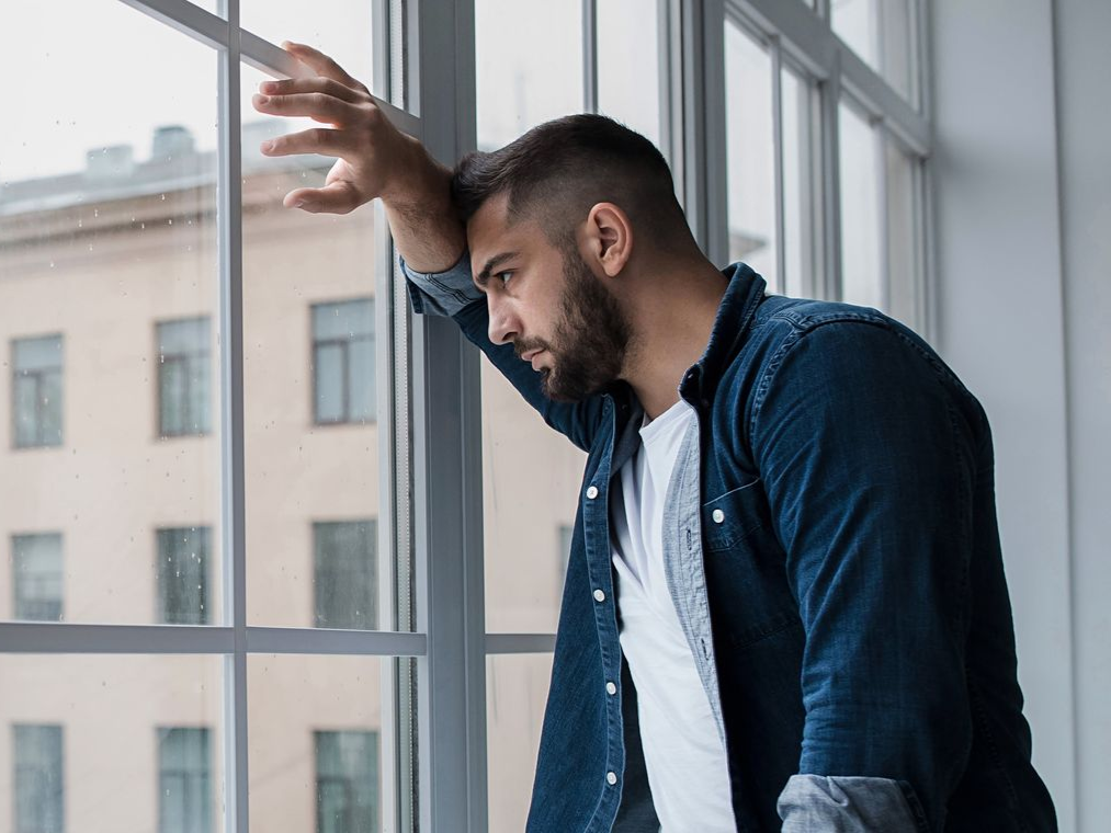 Man with a beard, leaning against a window, looking out with a concerned expression.