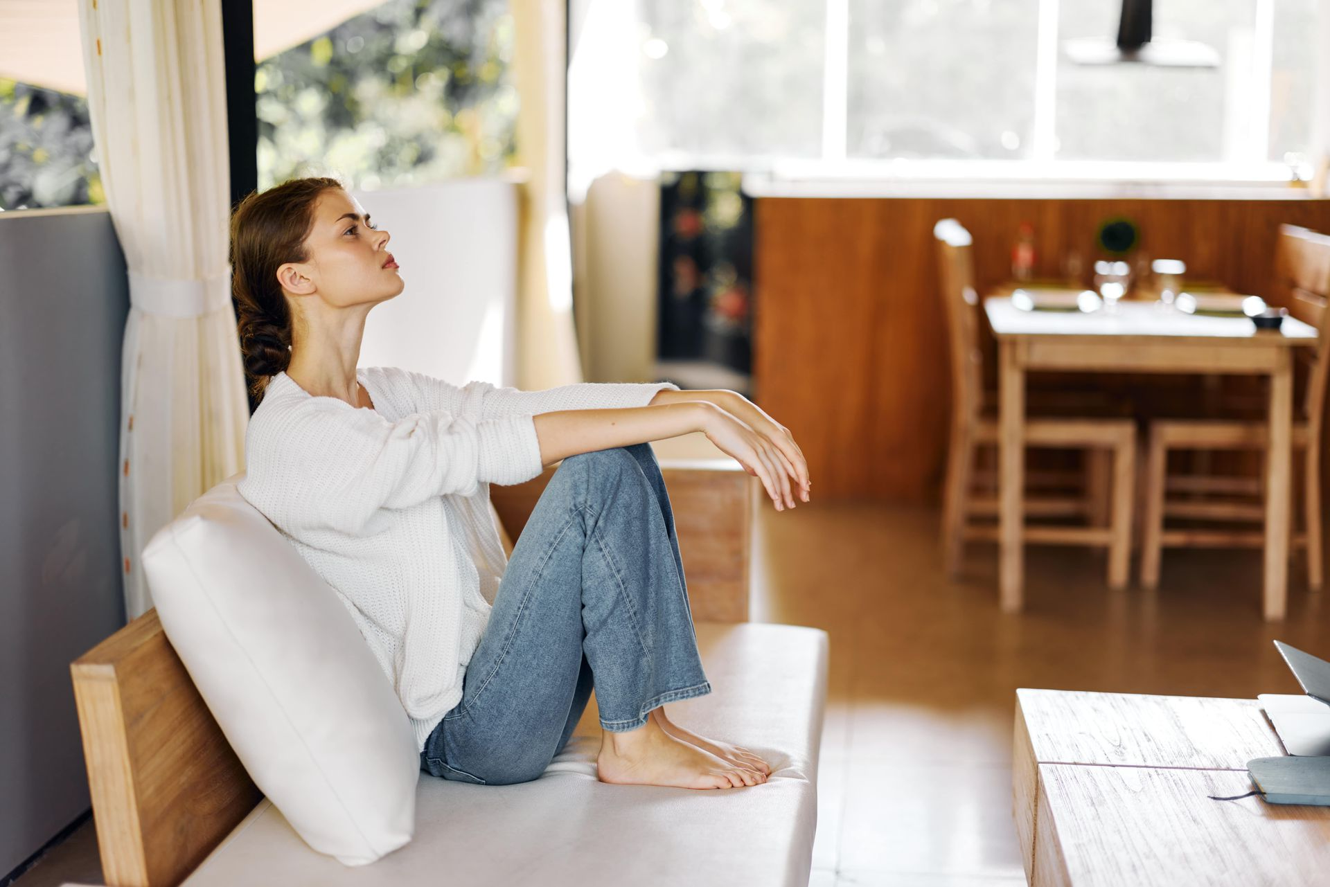 Woman sitting barefoot on a couch, eyes closed, indoors. Light-filled room with a table and kitchen in background.