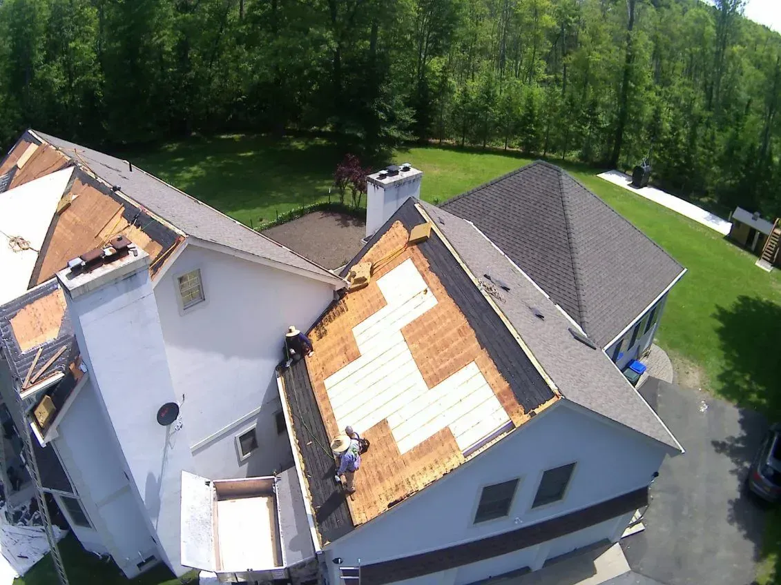 Roofers working on a residential roof, partially stripped of shingles, with a backdrop of trees and a sunny sky.