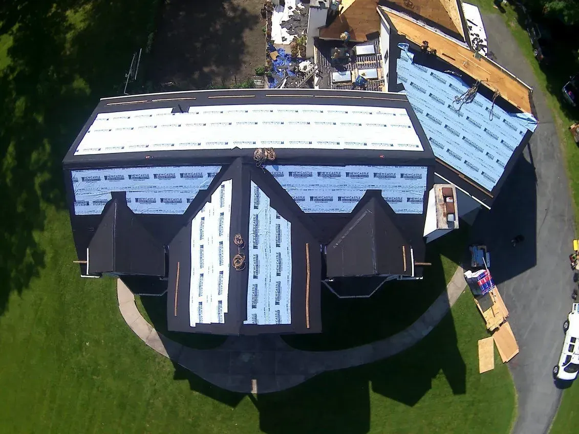 Overhead view of a house roof under construction, with new black shingles and blue tarp in progress, surrounded by green lawn.