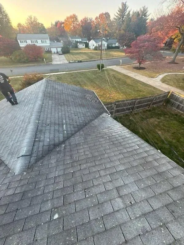 Overhead view of a house roof covered in gray shingles with a chimney, a lawn, and residential neighborhood in the background.